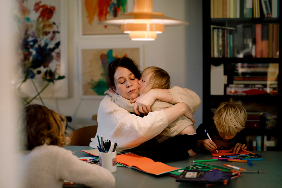 Mother hugging daughter while other children draw and color at table. 