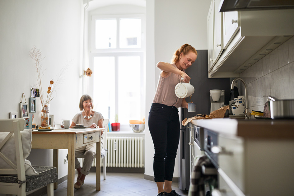 Middle-aged daughter brewing coffee for her elderly mother sitting at table in kitchen. 