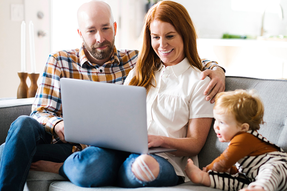 Husband and wife in their living room with their toddler looking at laptop. 