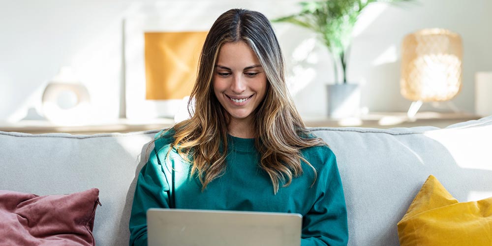 A smiling young woman sitting on a couch at home and working on a laptop computer