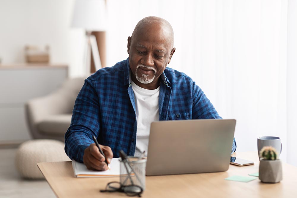 Elderly man using a laptop and taking notes with a pen and notebook