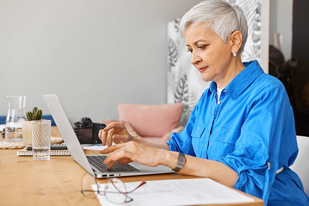 People, job, occupation, age and employment. Indoor image of beautiful gray haired female on retirement looking for remote work using portable computer. Mature woman photographer typing on laptop