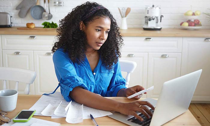 Indoor shot of attractive serious young dark skinned with wavy hair doing paperwork in kitchen, typing numbers in online form, holding paper bill, paying for gas, electricity and rent using laptop