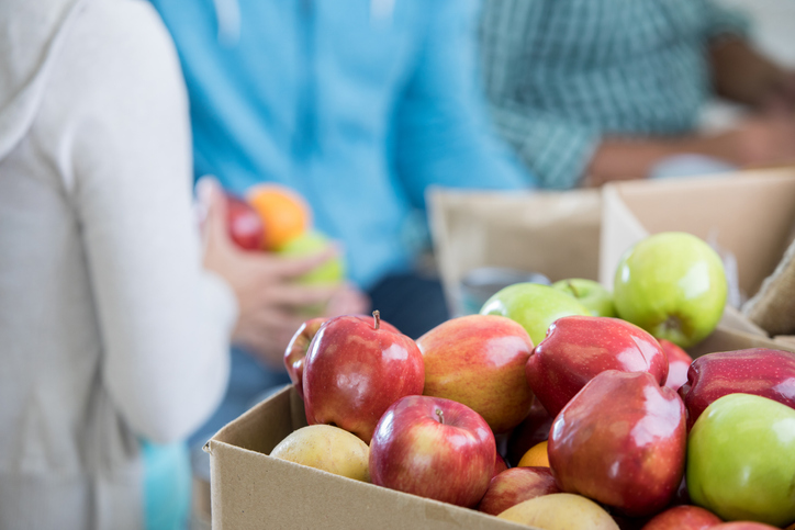 Close up of a box of red and green apples. People are in the background organizing the food that has been donated to a food bank.