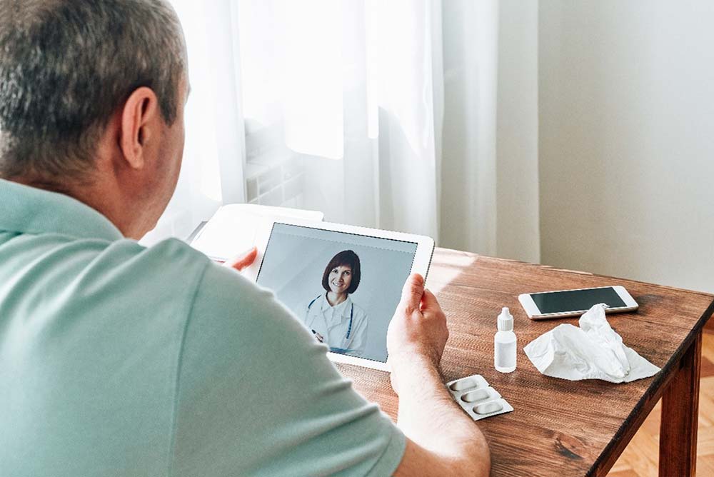 Older man holding iPad with doctor on screen for telehealth appointment.