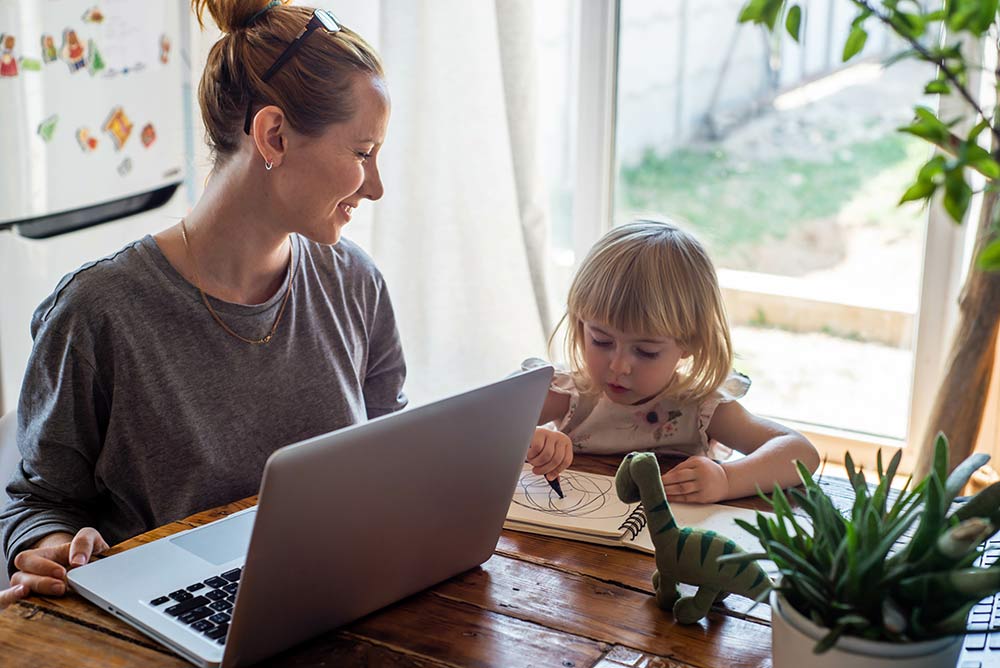 A mom at home using a laptop with young daughter at the table