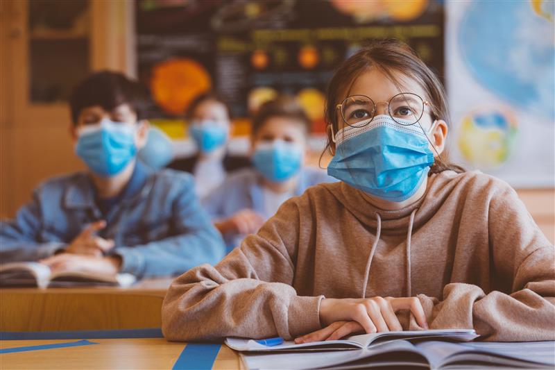Girl student wearing glasses and mask with book in middle school classroom.