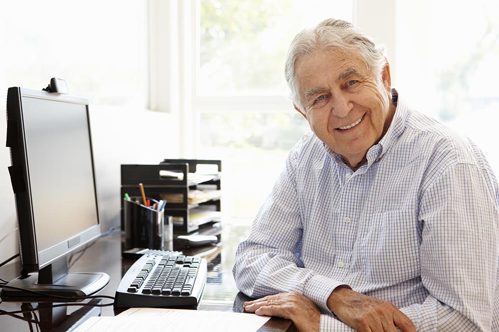 Smiling elderly man sits in front of a desktop computer.
