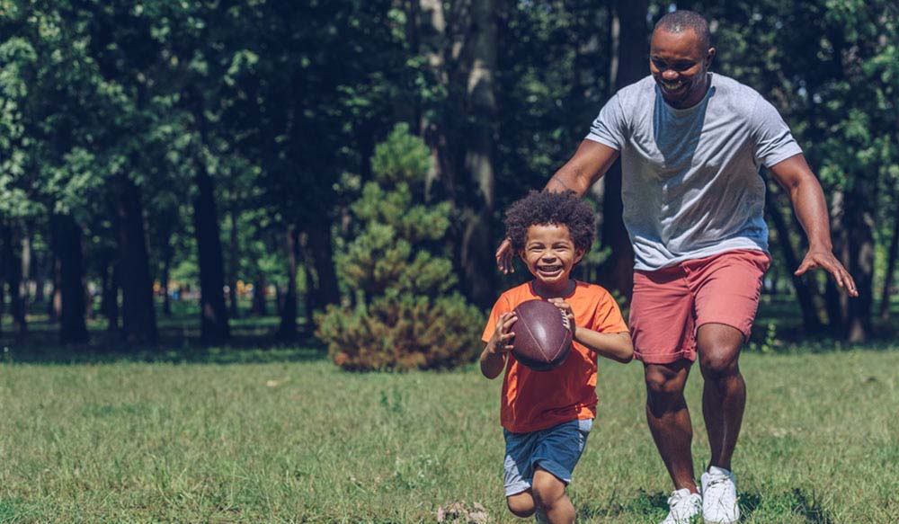 cute african american boy running with rugby ball while having fun with father in park