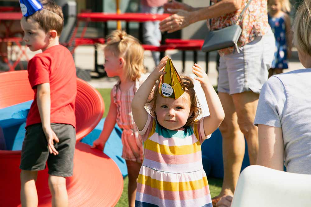 Young girl wearing a birthday hat with a BCBSND Caring Foundation sticker on the front, at the Red River Market Opening Day, sponsored by BCBSND Caring Foundation 