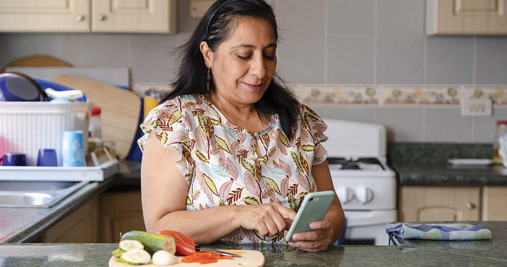 Hispanic mom searching recipes on her phone-mature woman preparing healthy and organic salad while checking her cell phone-housewife cooking while she looks at a phone