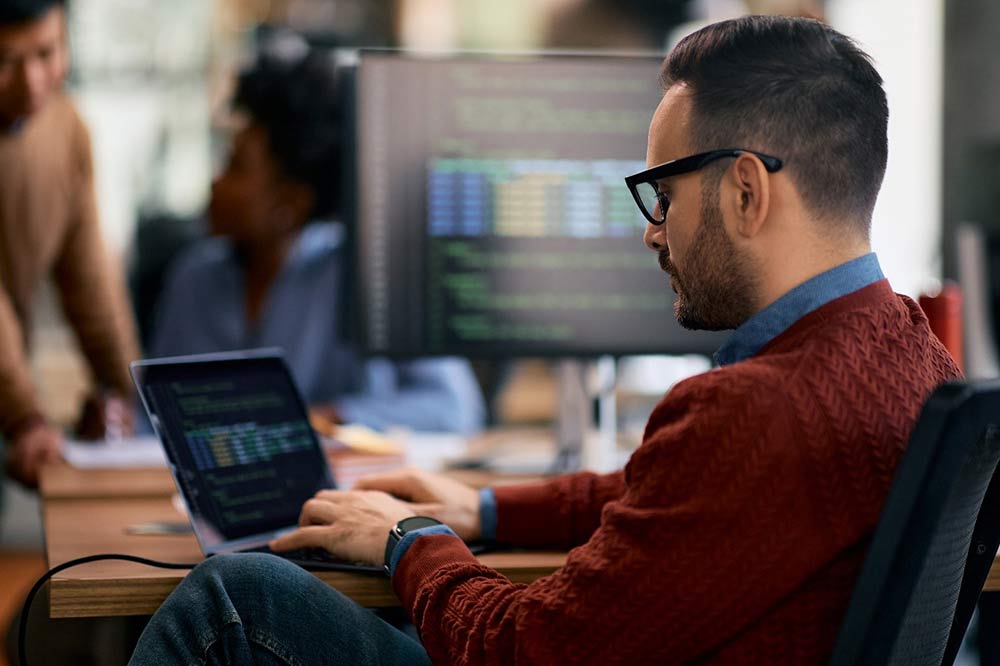 Man in business casual clothing sits at a desk and works on a laptop.